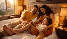 Une famille installée sur un lit dans une chambre chaleureusement éclairée, avec vue sur une ville par la fenêtre. Deux adultes sont assis contre la tête de lit, proches l’un de l’autre, tandis qu’un enfant est allongé et s’appuie sur eux. L’ambiance est détendue et conviviale, avec des lampes allumées créant une lumière douce.
