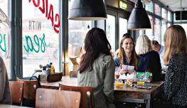 Un groupe de trois femmes discute autour d'une table de restaurant, avec des boissons sur la table et des luminaires noires au dessus de leur table. Elles sont contre une baie vitrée avec les inscriptions du restaurant dessus.