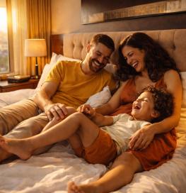 Une famille installée sur un lit dans une chambre chaleureusement éclairée, avec vue sur une ville par la fenêtre. Deux adultes sont assis contre la tête de lit, proches l’un de l’autre, tandis qu’un enfant est allongé et s’appuie sur eux. L’ambiance est détendue et conviviale, avec des lampes allumées créant une lumière douce.