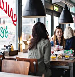Un groupe de trois femmes discute autour d'une table de restaurant, avec des boissons sur la table et des luminaires noires au dessus de leur table. Elles sont contre une baie vitrée avec les inscriptions du restaurant dessus.