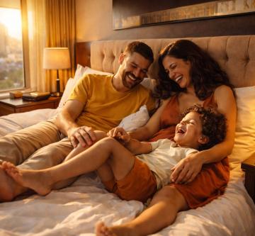 Une famille installée sur un lit dans une chambre chaleureusement éclairée, avec vue sur une ville par la fenêtre. Deux adultes sont assis contre la tête de lit, proches l’un de l’autre, tandis qu’un enfant est allongé et s’appuie sur eux. L’ambiance est détendue et conviviale, avec des lampes allumées créant une lumière douce.