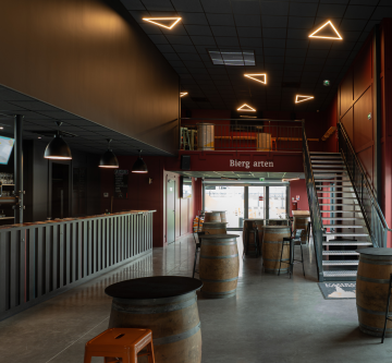 Espace intérieur de bar avec mezzanine, escalier métallique, tables tonneaux et plafond acoustique foncé