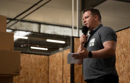 Intervenant s’exprimant au micro lors d’une conférence ou rencontre professionnelle, dans un lieu intérieur au design industriel avec panneaux en bois et éclairage suspendu.