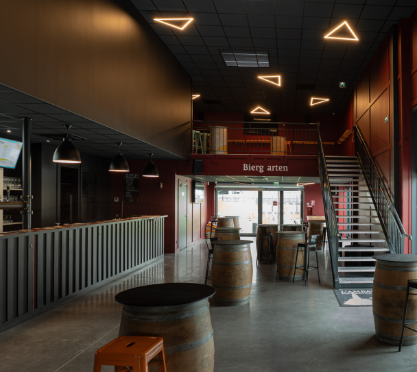 Espace intérieur de bar avec mezzanine, escalier métallique, tables tonneaux et plafond acoustique foncé